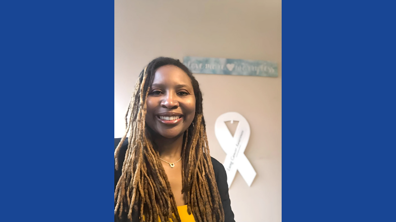 A woman stands in front of a white cutout ribbon on a cream colored wall.